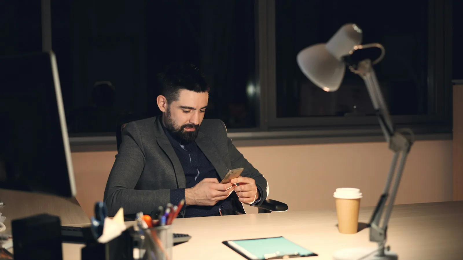 A man working late at a lamp-lit desk, looking at his phone