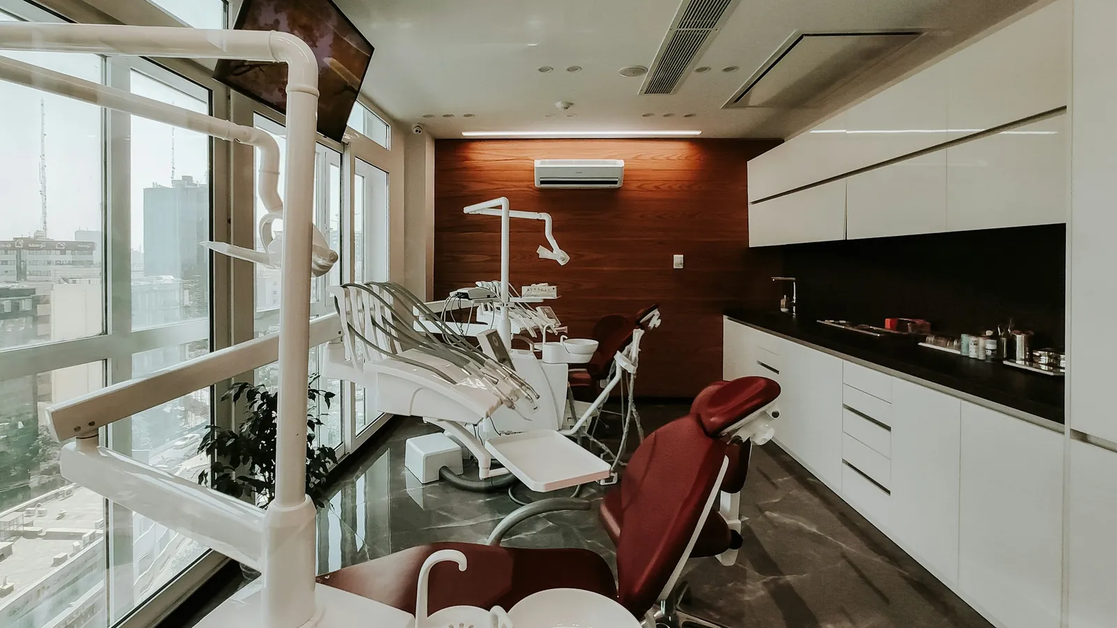 An empty dental treatment room with a red patient chair by the windows