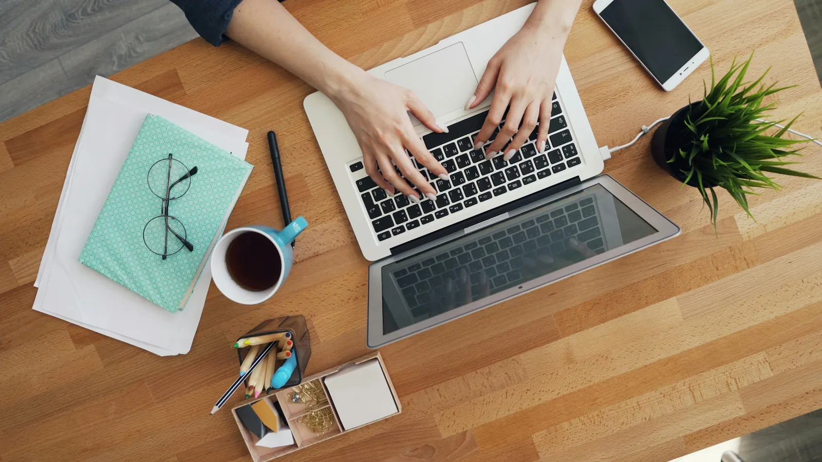 Overhead view of hands typing on a laptop on a wooden desk, with a phone, glasses and a cup of tea