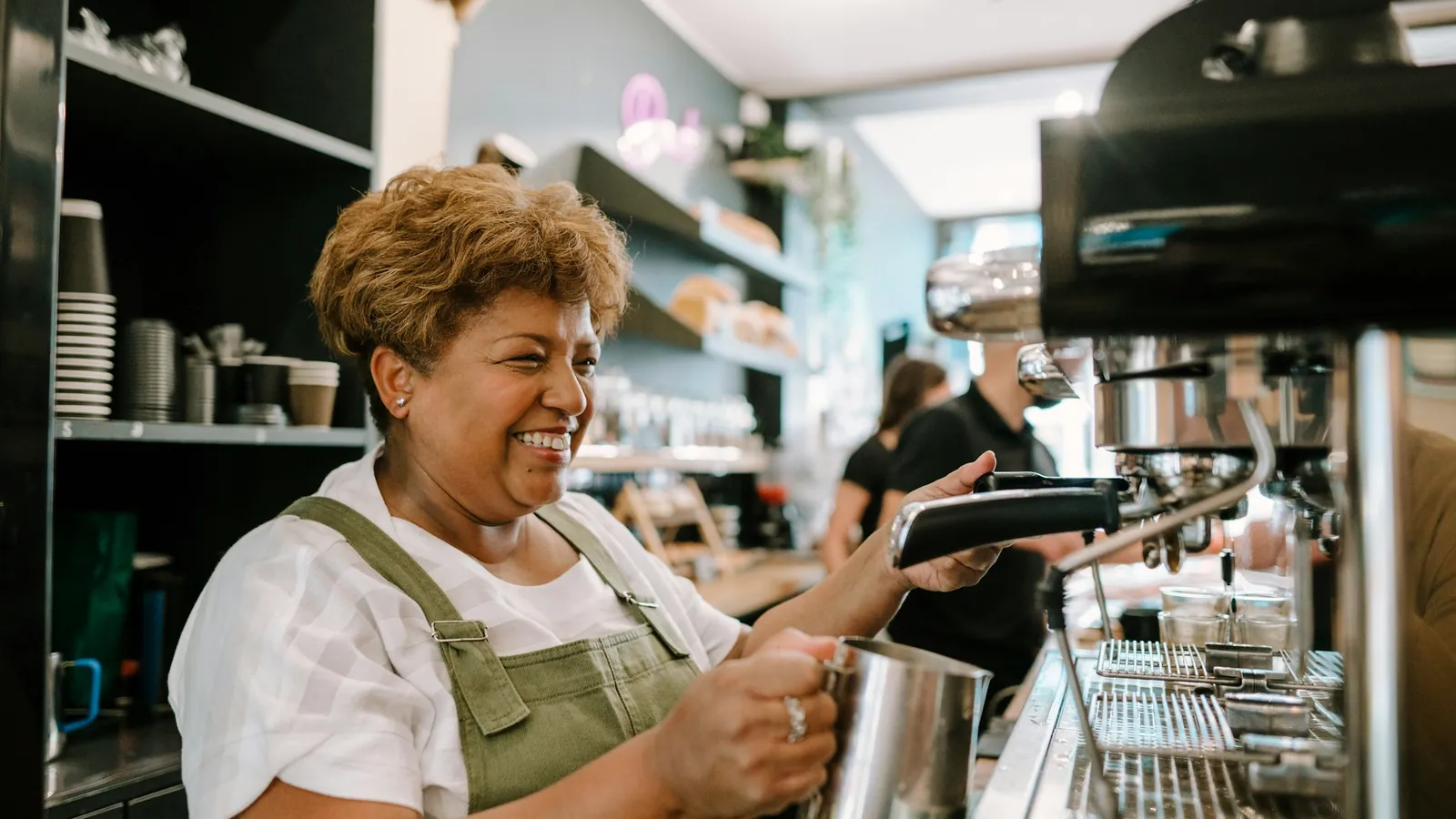A smiling woman steaming milk at a café's espresso machine