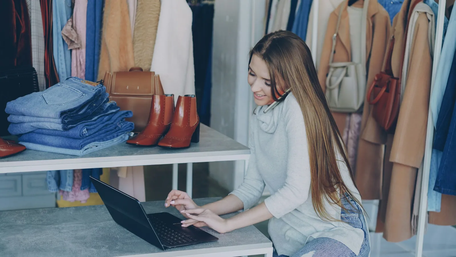 A woman working on a laptop and phone in a clothing shop, between racks of clothes