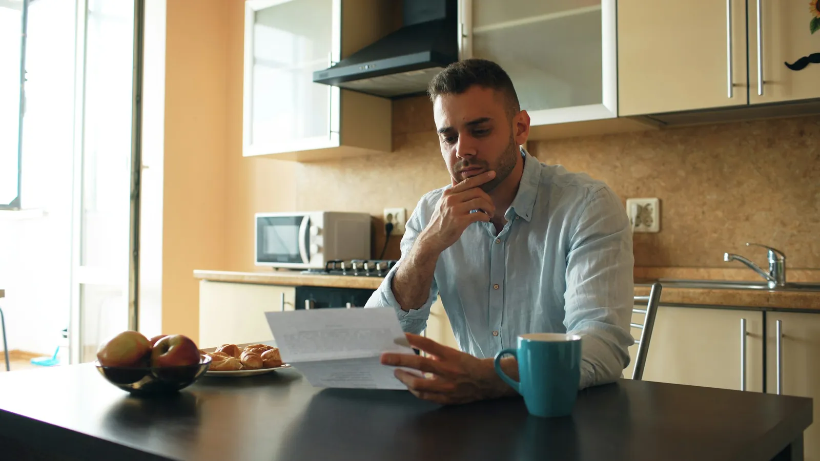 A man at a kitchen table reading a printed document, with a mug of coffee beside him